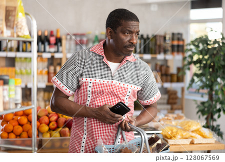 In store, African man with phone in hands looking for something on shelf of showcase In store, African man with phone in hands looking for something on shelf of showcase 128067569