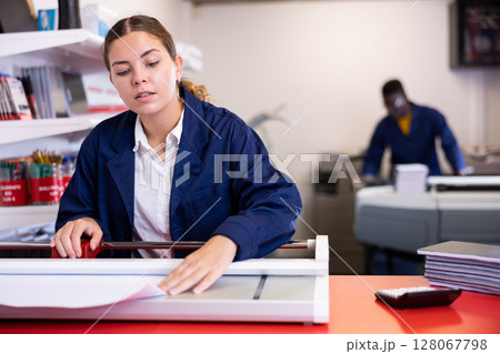 Serious young woman in a blue uniform using paper cutter on the table with planners and calculator in the printer house Serious young woman in a blue uniform using paper cutter on the table with planners and calculator in the printer house 128067798