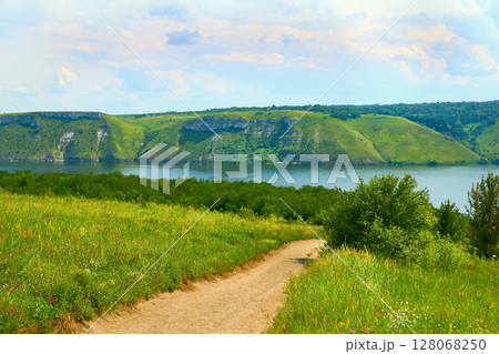 A rural field road leading to a calm river with hilly banks 128068250
