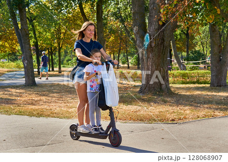 Cheerful mother and daughter riding electric scooter in summer park Cheerful mother and daughter riding electric scooter in summer park 128068907