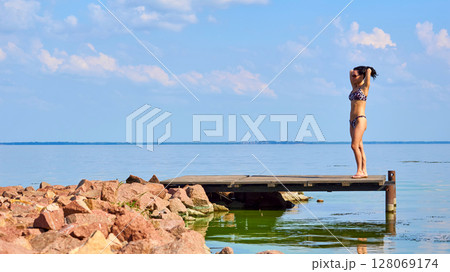 Young pretty woman in swimsuit standing on pier on shore Young pretty woman in swimsuit standing on pier on shore 128069174