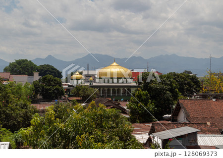 Golden dome of a mosque rises above traditional houses and trees with mountains in the background 128069373