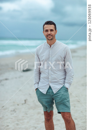 Handsome man in shirt relax on the beach. Laughing beach guy in shorts and shirt in cloudy day on the coast 128069938