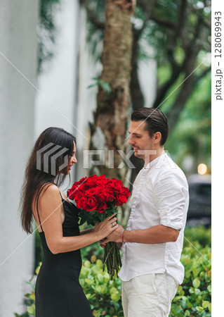 Young man gives his girlfriend a bouquet of red roses on Valentine's Day. The girl is wearing an elegant black dress. Valentine's Day Flowers Young man gives his girlfriend a bouquet of red roses on Valentine's Day. The girl is wearing an elegant black dress. Valentine's Day Flowers 128069943