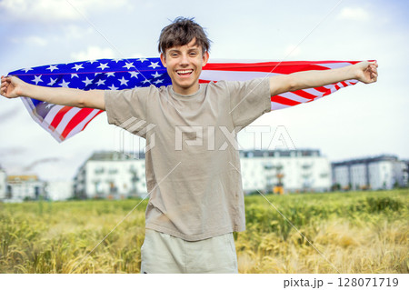 Happy and cheerful teenage boy with arms spread wide waving American flag in a field  128071719
