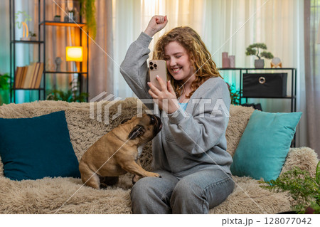 Young woman reacts happily to good news on smartphone with pug dog on home sofa smiling with emotion 128077042