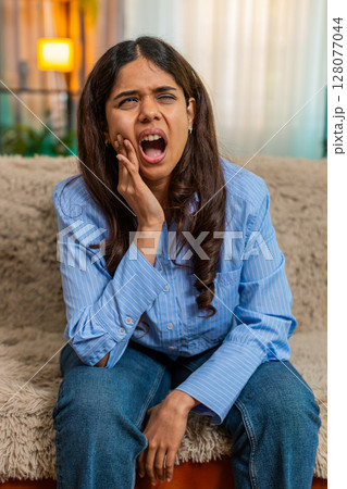 Indian woman holding cheek from sharp toothache pain on home sofa, showing discomfort and tension 128077044