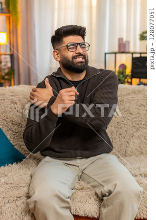 Indian man sitting at home holds shoulder with discomfort, reacting to physical pain and tension 128077051