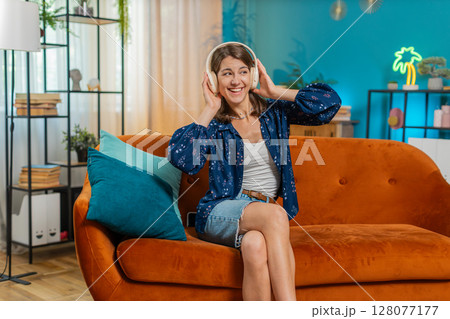 Joyful young woman enjoying listening to music through wireless headphones dancing at home room sofa Joyful young woman enjoying listening to music through wireless headphones dancing at home room sofa 128077177