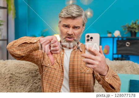 Mature man sitting on sofa with smartphone showing thumbs down, expressing disappointment at home Mature man sitting on sofa with smartphone showing thumbs down, expressing disappointment at home 128077178