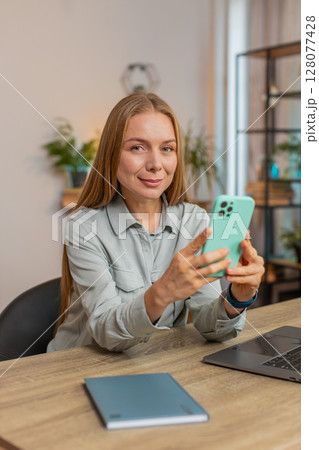 Happy female freelancer sitting at desk using smartphone texting smiling in living room at home 128077428