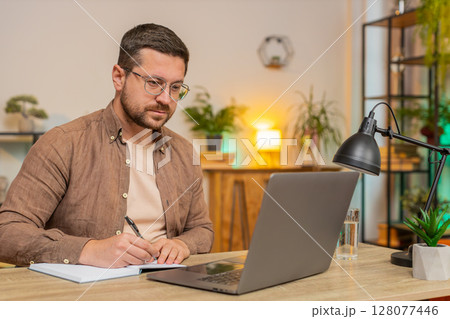 Young freelancer writing down notes while attending online meeting on laptop at home office desk 128077446