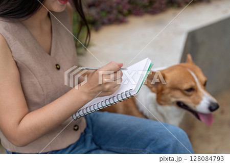 Note-taking and Pet Interaction. Woman writing notes with her corgi dog beside her. 128084793