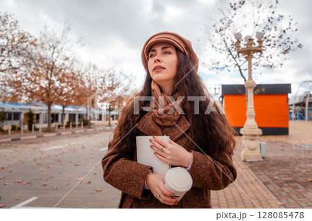 Portrait of young woman in a beret and coat is walking around the city with a book and drinking coffee. City leisure. Bottom view Portrait of young woman in a beret and coat is walking around the city with a book and drinking coffee. City leisure. Bottom view 128085478