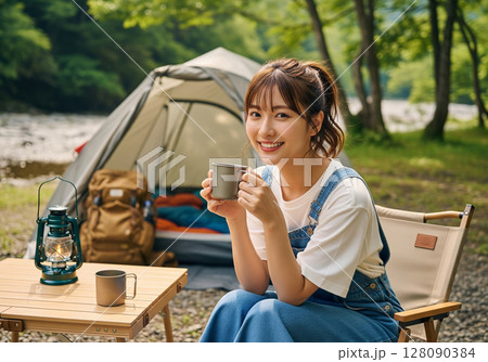 キャンプ場で笑顔、白Tシャツとオーバーオールを着てマグカップを持つ女性モデル 128090384