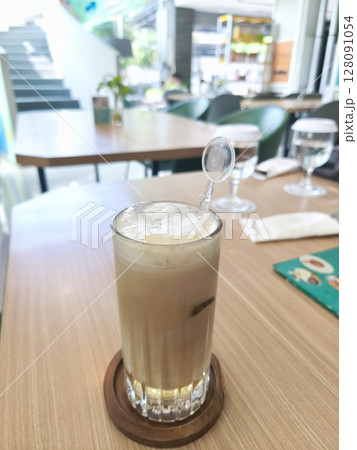 An iced coffee drink with froth and a stirrer sits on a wooden coaster, next to a glass of water, on a wooden table. An iced coffee drink with froth and a stirrer sits on a wooden coaster, next to a glass of water, on a wooden table. 128091054