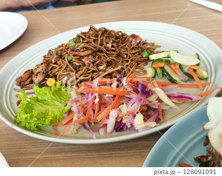 A plate of Indonesian Mie Goreng or fried noodles with chicken, vegetables, pickled cucumber and carrot, and fresh salad, on a wooden table in Surabaya. A plate of Indonesian Mie Goreng or fried noodles with chicken, vegetables, pickled cucumber and carrot, and fresh salad, on a wooden table in Surabaya. 128091091