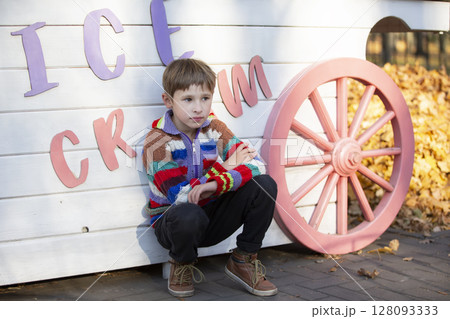 A little boy sits in the park near the wall with the inscription "Ice Cream". The child went out for a walk. 128093333