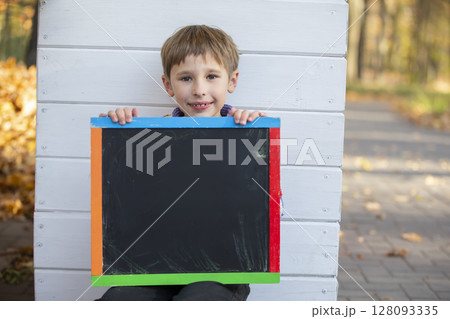 A little boy sits in the park near the wall and holds an empty black board for the inscription. A little boy sits in the park near the wall and holds an empty black board for the inscription. 128093335