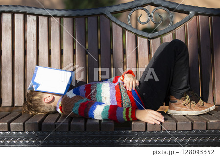 A little boy in the park lies on a bench and his face is covered with a book. 128093352