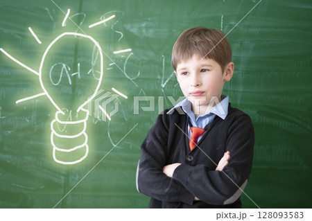 Thinking over an idea. Child student in class. Schoolboy at the green chalkboard with a painted light bulb. Primary school education. 128093583