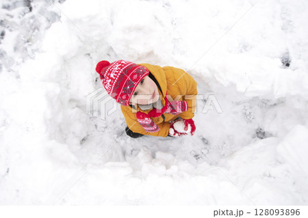 Funny little boy in colorful clothes playing outdoors during a snowfall. The child looks up from the snow pit. 128093896