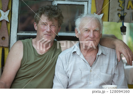 An adult son and an old man father are hugging and looking at the camera. 128094160