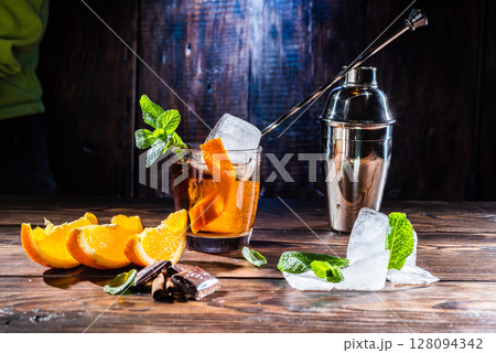 Cocktail with ice and orange. Cocktail spoon in a glass. Orange slices and chocolate in the foreground. Cocktail shaker. 128094342