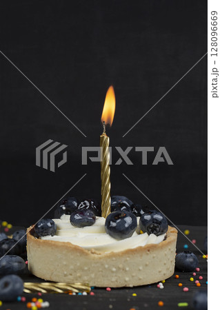 On a dark background, a cake with a burning candle sprinkled with black berries. 128096669