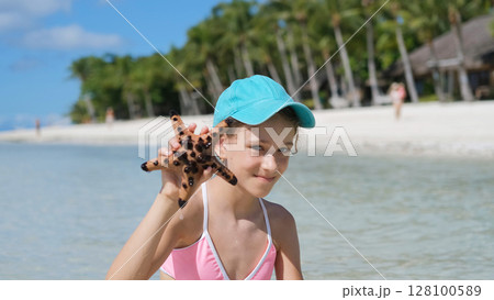 Girl wades in ocean holding starfish near tropical beach with white sands Girl wades in ocean holding starfish near tropical beach with white sands 128100589
