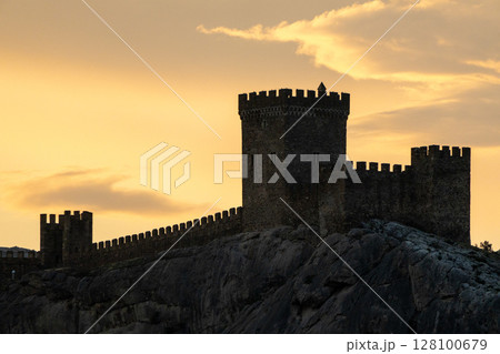Castle Genoese Sunset Silhouette of a medieval fortress with towers and battlements on a rocky hill, against a warm sunset sky with light clouds. 128100679