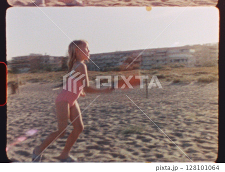 Young girl energetically playing beach tennis, hitting ball with racket on sandy coastal landscape, capturing joyful summer vacation moment near sparkling sea waters - filmed on super 8 mm film 128101064