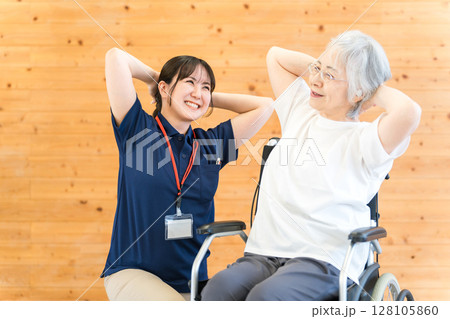 リハビリで体操をする車椅子に乗ったシニア女性と理学療法士・作業療法士の女性スタッフ 128105860