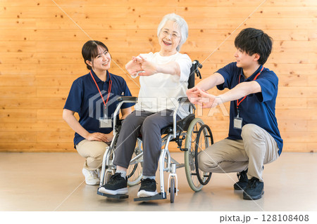 リハビリで体操をする車椅子に乗ったシニア女性と理学療法士・作業療法士の男女スタッフ 128108408