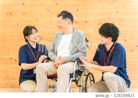 車椅子のシニア男性と介護士・ケアマネージャーの男女スタッフ（認知症・リハビリ） 128108621