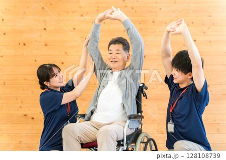 リハビリで体操をする車椅子に乗ったシニア男性と理学療法士・作業療法士の男女スタッフ リハビリで体操をする車椅子に乗ったシニア男性と理学療法士・作業療法士の男女スタッフ 128108739