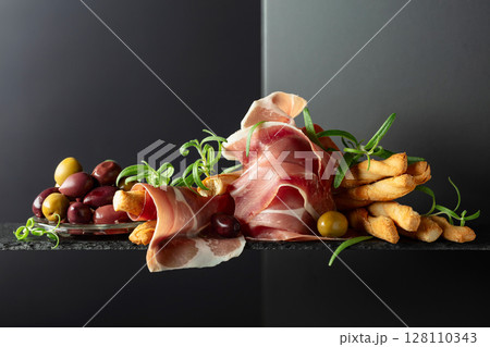 Prosciutto with bread sticks, olives, and rosemary on a black background. Prosciutto with bread sticks, olives, and rosemary on a black background. 128110343