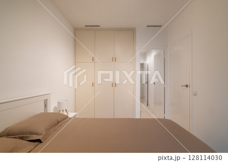 Minimalist bedroom view with taupe bedding, white built-in wardrobe and clean neutral walls Minimalist bedroom view with taupe bedding, white built-in wardrobe and clean neutral walls 128114030