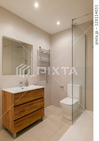 Bathroom with beige tiled walls, wood vanity with white sink, glass shower and chrome fixtures 128114031