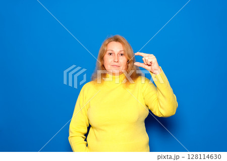 Caucasian woman in her 40s wearing a yellow sweater, smiling and confident, gesturing with her hand, making a small sign with her fingers, looking at the camera. Isolated over blue background. Caucasian woman in her 40s wearing a yellow sweater, smiling and confident, gesturing with her hand, making a small sign with her fingers, looking at the camera. Isolated over blue background. 128114630