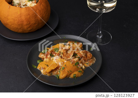 Whole baked pumpkin with tiger prawns, melted cheese, tomatoes and a glass of white wine. Traditional dish of Brazil, home cooking. Selective focus, close-up, black background. 128123927