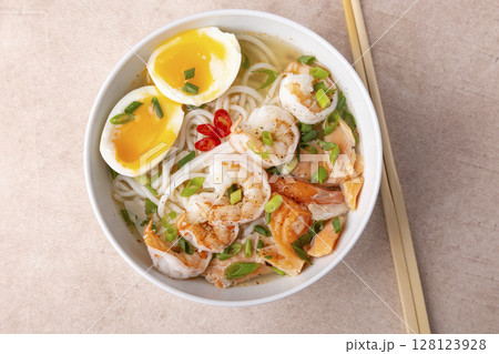 Asian dish with broth, noodles, tiger shrimp, fish, egg and green onions. Traditional Asian soup in a white deep plate. Selective focus, close-up. Asian dish with broth, noodles, tiger shrimp, fish, egg and green onions. Traditional Asian soup in a white deep plate. Selective focus, close-up. 128123928