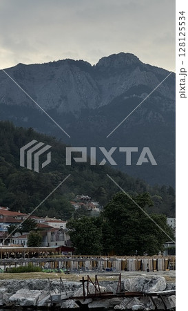 View of a small Greek town at the base of a forested mountain under an overcast sky, ideal for travel and landscape themes. 128125534