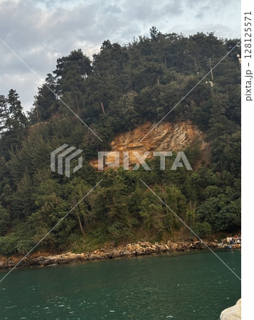 A tree-covered cliff with exposed rock rises above turquoise water, representing coastal wilderness and Mediterranean landscapes. 128125571