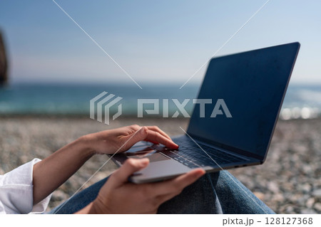 Laptop Beach Working: Woman types outdoors on laptop by sea during daytime for remote work. Laptop Beach Working: Woman types outdoors on laptop by sea during daytime for remote work. 128127368