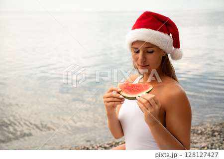 A woman is eating a watermelon while wearing a red santa hat. Scene is lighthearted and festive, as the woman is enjoying a refreshing snack on a sunny day. A woman is eating a watermelon while wearing a red santa hat. Scene is lighthearted and festive, as the woman is enjoying a refreshing snack on a sunny day. 128127427