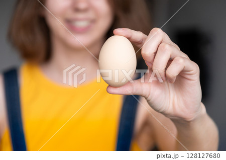 One chicken egg is held in a woman's hand. Concept of demonstrating sizes. Chicken egg. Hand holds a chicken egg. Girl's hand shows an egg on the background of itself One chicken egg is held in a woman's hand. Concept of demonstrating sizes. Chicken egg. Hand holds a chicken egg. Girl's hand shows an egg on the background of itself 128127680