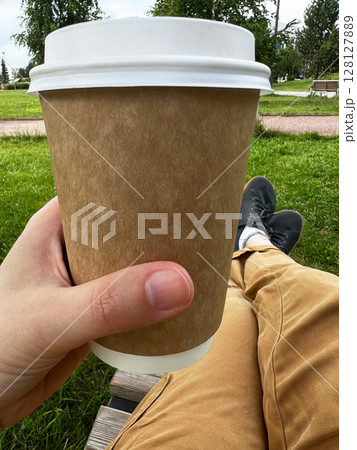 Hand holding a disposable coffee cup with a brown sleeve, resting on a wooden bench in a green park, showcasing a relaxed outdoor lifestyle and enjoyment of beverages 128127889
