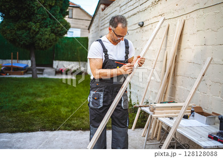 Mature male carpenter measuring wooden plank outdoors 128128088