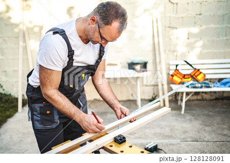 Mature male carpenter measuring wooden plank outdoors Mature male carpenter measuring wooden plank outdoors 128128091
